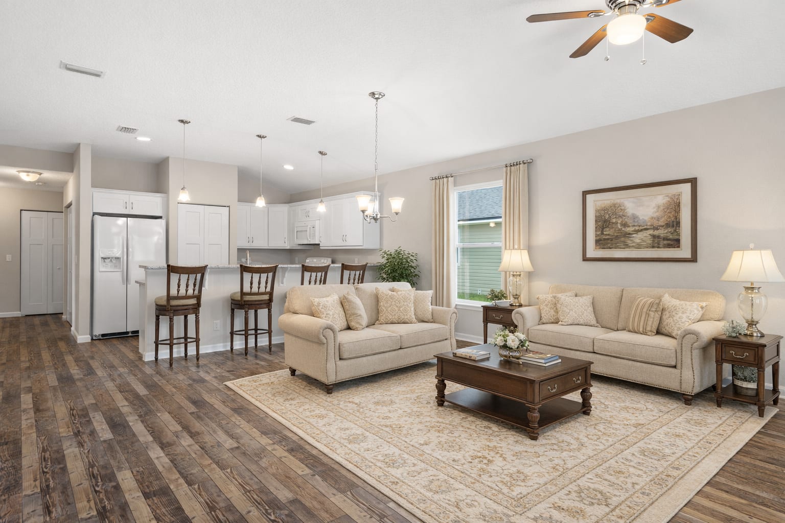 Same room with virtual staging applied: two beige sofas, a wood coffee table, table lamps, framed art, an area rug. Same kitchen, same fan, same floor, same window. The room is now furnished.