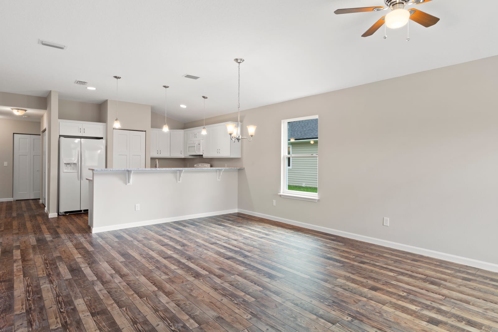 Empty open-plan great room: open kitchen with white cabinets and a breakfast bar, then a wide unfurnished living area with dark wood floors, a ceiling fan, and a single window letting in daylight.