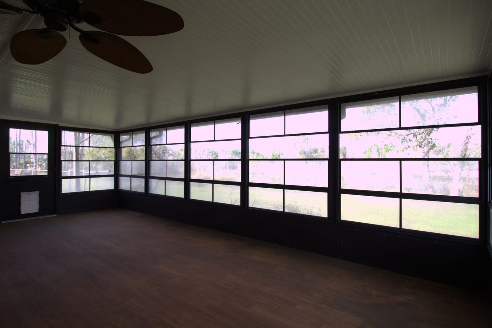 Empty enclosed sunroom with six-panel windows, dark wood floor, wicker ceiling-fan blades. Window views are blown out from the bright outside light.