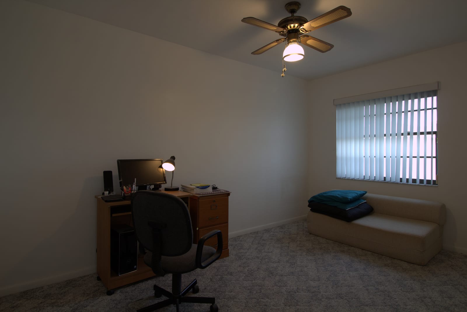 Cluttered home office with brown carpet, vertical-blind window, ceiling fan-light, small desk with monitor and lamp in active use, and a futon piled with bedding. Reads as multi-use and dim.